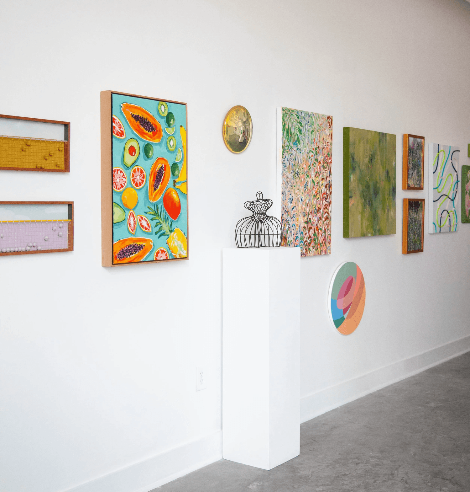 Gallery wall featuring colorful paintings, mixed media art pieces, fruit motifs, and abstract designs displayed in a bright minimalist gallery space